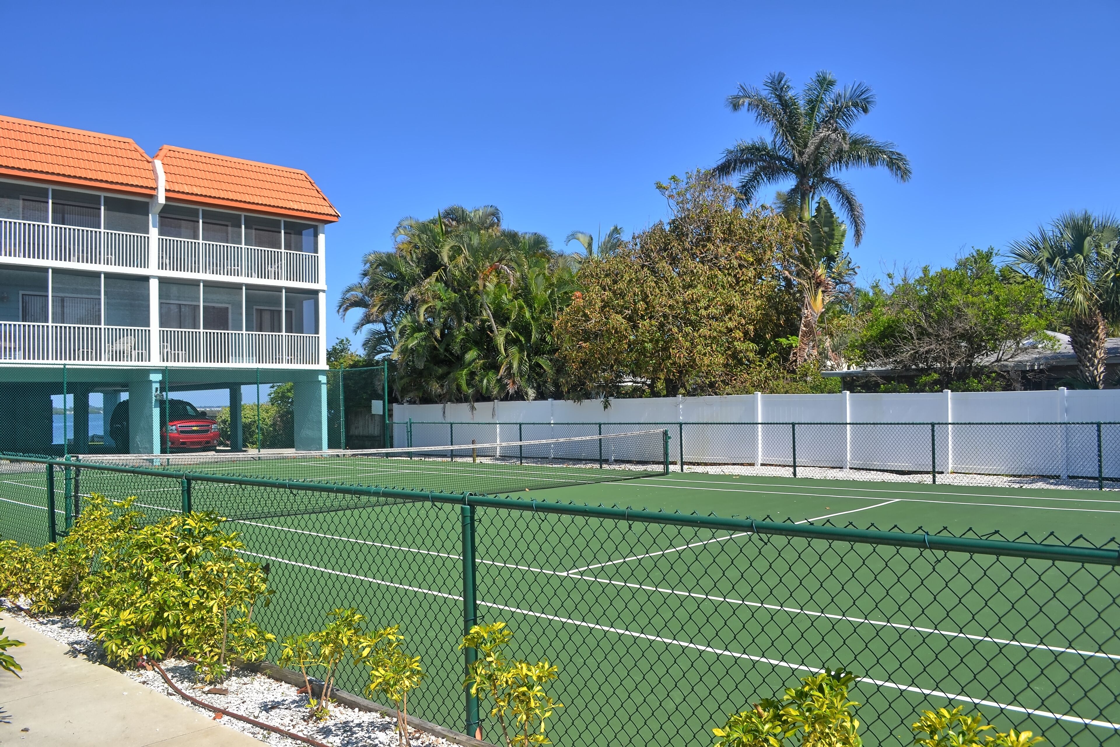 A tennis court with a green fence, a red car, and a building with a red roof.