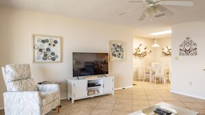 Una sala de estar con televisor, un mueble blanco para equipo audiovisual, un sillón estampado y una zona de comedor con candelabro.