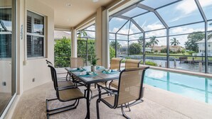 A covered outdoor area with a pool, a table with chairs, and large windows offering a view of the surrounding area. - June Court | Waterfront 4BR w/ Pool, Dock, Open Layout & Near Beach (Marco Island)