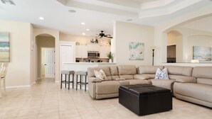 A spacious living room with a beige sectional sofa, a black ottoman, and a kitchen in the background. - June Court | Waterfront 4BR w/ Pool, Dock, Open Layout & Near Beach (Marco Island)