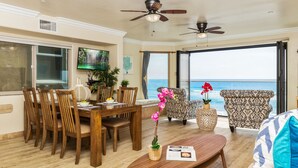 A spacious living area with a dining table, chairs, and a large window offering a view of the ocean. There are ceiling fans, a television, and a potted plant. - Dreamy Coastal Haven Escape P819-5 (Oceanside)