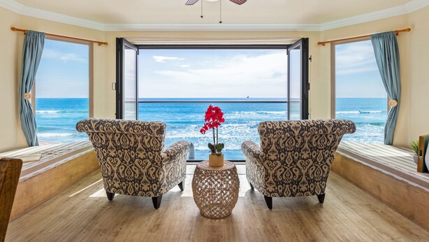 A room with a large window overlooking the ocean, two patterned armchairs, a small round table with a vase of red flowers, and a ceiling fan. - Dreamy Coastal Haven Escape P819-5 (Oceanside)