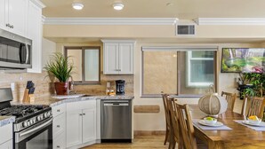 A modern kitchen with white cabinets, a stainless steel stove, and a dishwasher. There is a dining area with a wooden table and chairs, and a large window providing natural light. - Dreamy Coastal Haven Escape P819-5 (Oceanside)