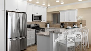 A modern kitchen with white cabinets, stainless steel appliances, a granite countertop, and a bar area with white chairs. - Ultimate Serene Escape P819-6 (Oceanside)