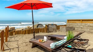 A beachside lounge with a red umbrella, wicker lounge chairs, and a table with snacks.