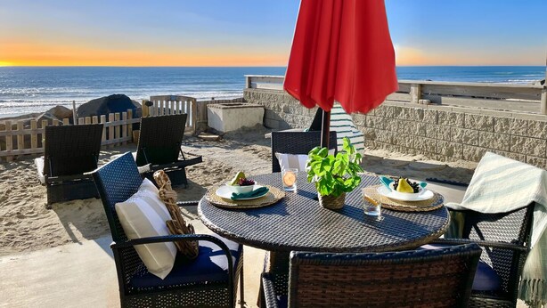 A beachside patio with wicker furniture, a red umbrella, and a table set with plates and a potted plant.