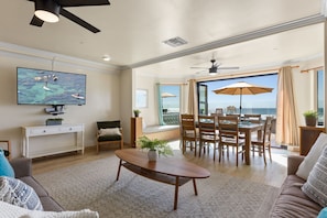 A spacious living area with a large TV, a dining table with chairs, a sofa, and a view of the ocean.