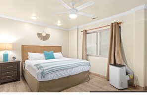 Ein Schlafzimmer mit Bett, Nachttisch, Deckenventilator, Fenster mit Vorhängen und Heizung.