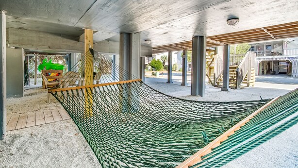 A hammock under a wooden pergola with a concrete ceiling, surrounded by a sandy area and a building with a wooden deck. - Tip Ellen on Pawleys Island - Second Row, Pet Friendly (Pawleys Island)