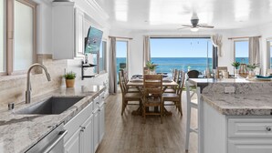 A kitchen with a view of the ocean, a dining area with wooden chairs, and a large island with a sink and countertop. - Spacious Beachfront Family Retreat - 819-4 (Oceanside)