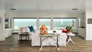 Uma sala de estar ampla com uma grande janela com vista para o oceano, um sofá branco, uma pequena mesa com um candeeiro e uma planta em vaso.