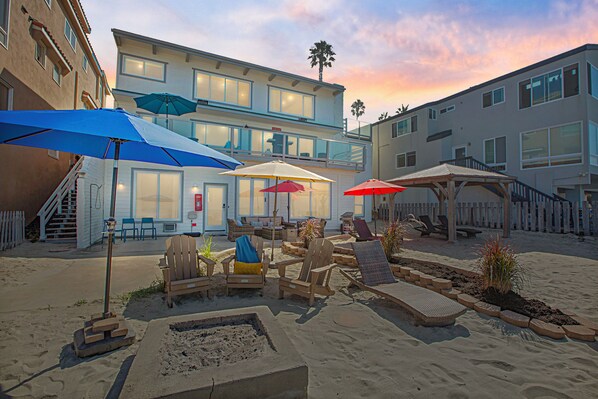 A beachfront property with outdoor seating, umbrellas, and a sandy patio area.