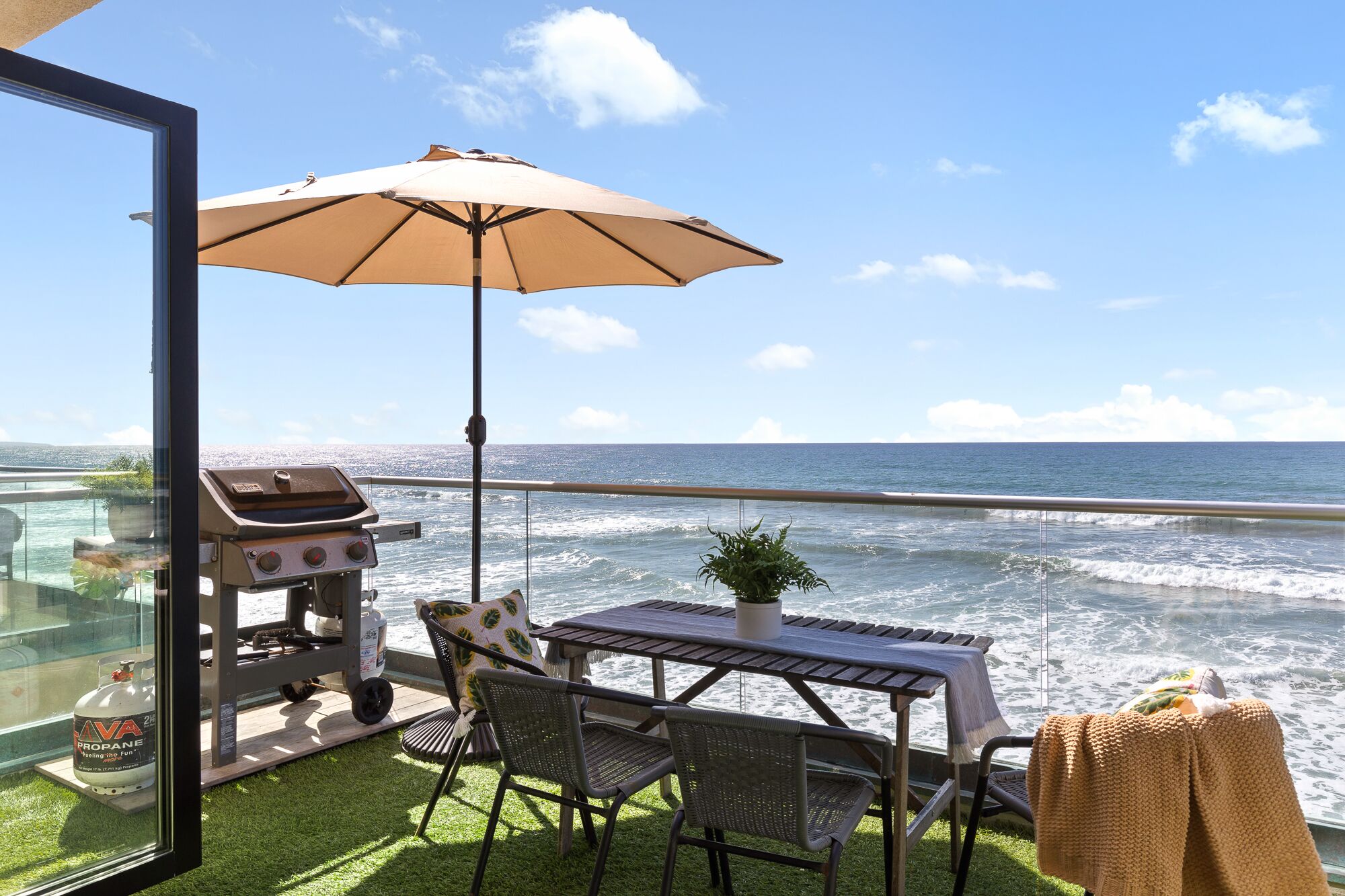 En udendørs patio med grill, bord, stole og parasol med udsigt over havet.