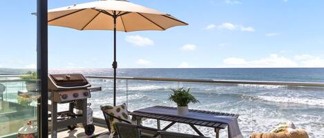En udendørs patio med grill, bord, stole og parasol med udsigt over havet.