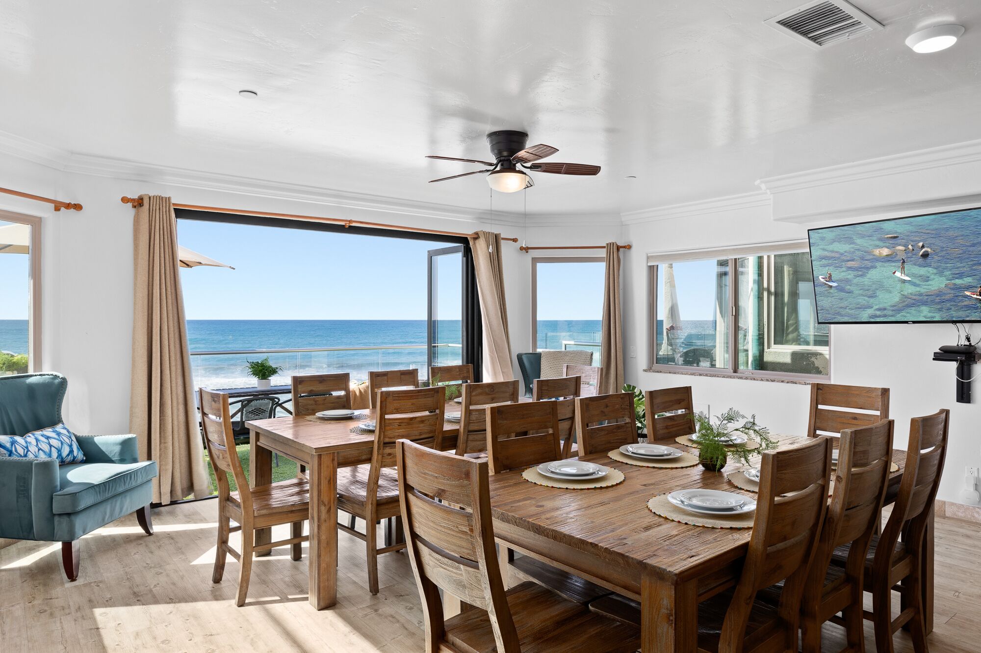 A dining area with a wooden table and chairs, large windows with ocean views, a ceiling fan, and a mounted TV.