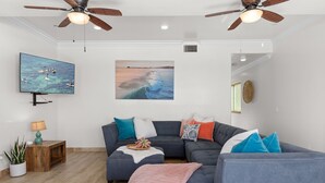 Una sala de estar con un sofá seccional, un televisor, un ventilador de techo y un cuadro con una escena de playa.