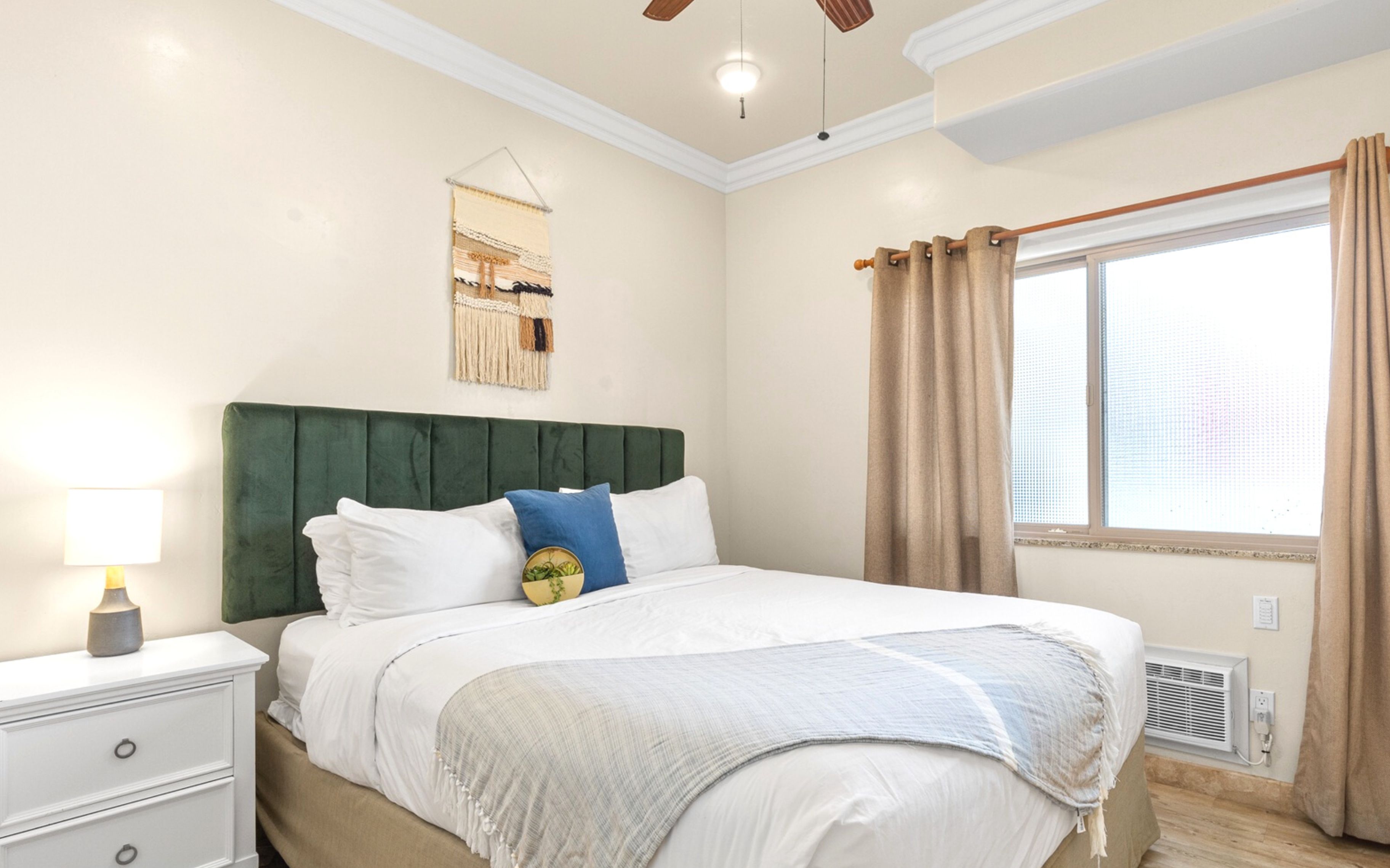 A bedroom with a green upholstered headboard, white bedding, a bedside lamp, and a window with curtains.
