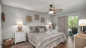 Un dormitorio con cama, dos mesitas de noche, ventilador de techo, ventana con cortinas y vista a zonas verdes.