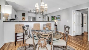 Un comedor con mesa de vidrio, sillas de mimbre, una araña y una cocina al fondo.