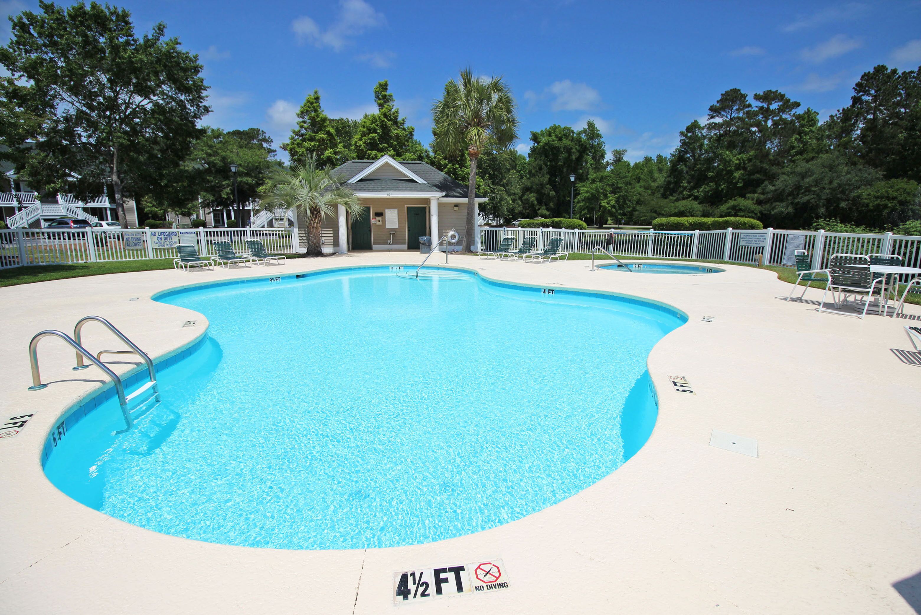 Une piscine dont la partie la moins profonde est marquée à 4 pieds et demi, entourée de chaises longues et d’un petit bâtiment, probablement un vestiaire ou des toilettes, avec une clôture blanche et des arbres en arrière-plan.