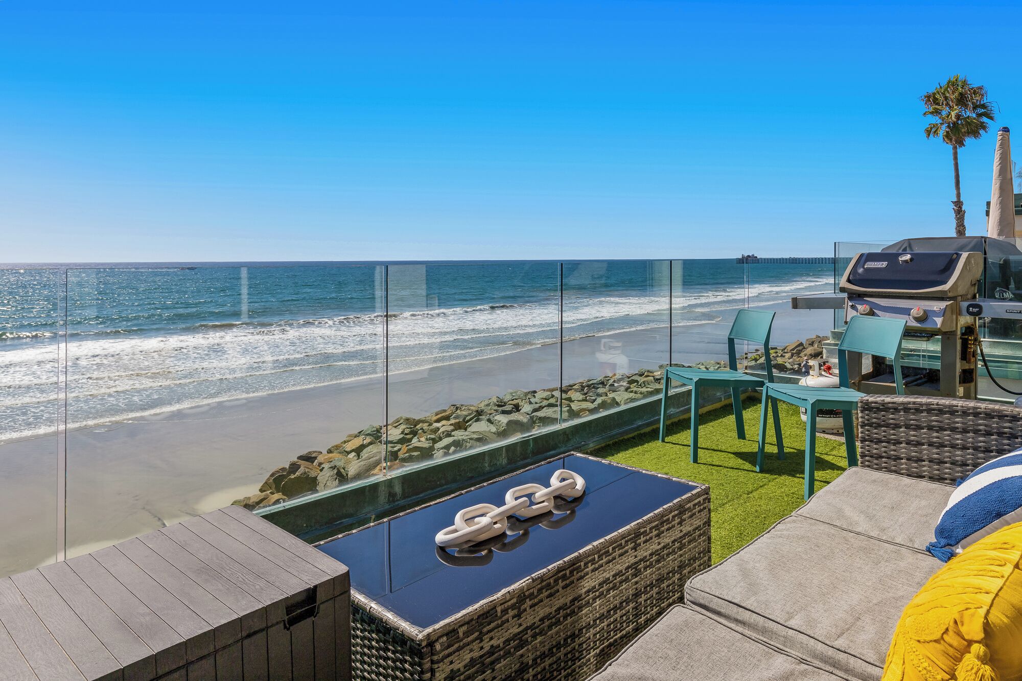 A balcony with a glass barrier overlooking a beach, a grill, and outdoor seating.