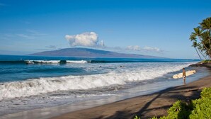 Une plage avec des vagues, une personne tenant une planche de surf et une montagne en arrière-plan.