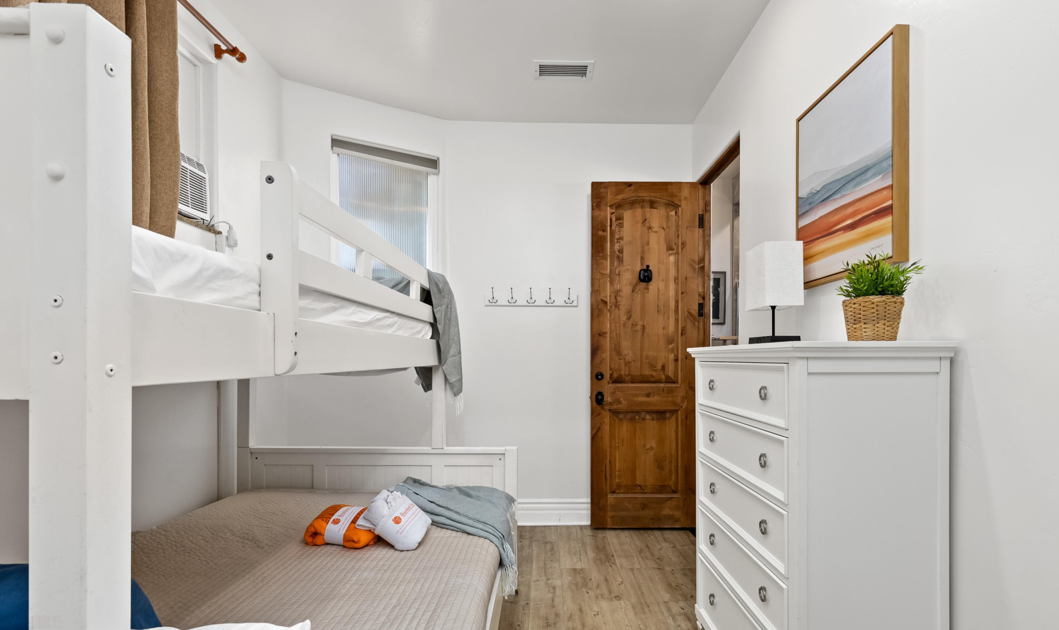 Una cama litera con cabecera de madera, una mesita de noche con lámpara, un aparador, un perchero y una puerta que da a otra habitación.
