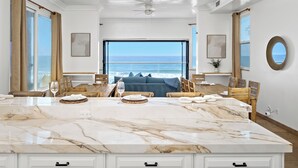 A spacious dining area with a large marble island, wooden chairs, and a view of the ocean. - Stunning Waterfront Oasis P823-1 (Oceanside)