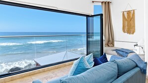 A modern living room with a large glass window overlooking the ocean, a beige sofa with blue cushions, and a macrame wall hanging. - Stunning Waterfront Oasis P823-1 (Oceanside)