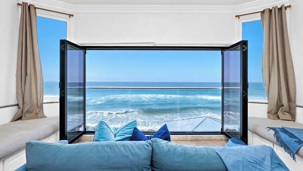 A room with a large window overlooking the ocean, a blue sofa with cushions, and beige curtains. - Stunning Waterfront Oasis P823-1 (Oceanside)
