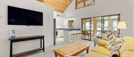 A modern living room with a flat-screen TV, wooden furniture, a kitchen in the background, and large windows offering a view.