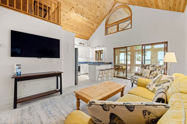 Un salón moderno con televisor de pantalla plana, muebles de madera, una cocina al fondo y ventanas amplias con vistas.