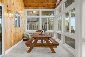 Un deck de madera con una mesa de picnic y bancos, ventanas amplias y un cartel con normas de la casa.