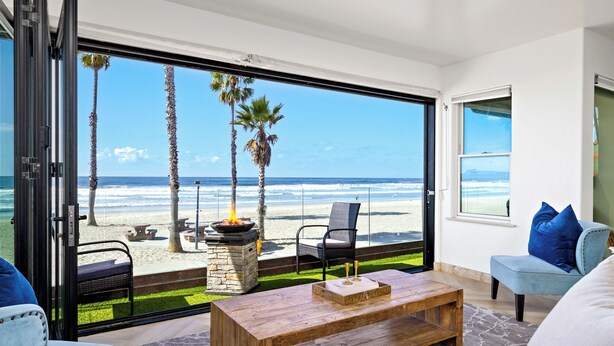A modern living room with a large glass door leading to a beach view, a wooden coffee table, and blue upholstered chairs. - Beachfront Bliss - Panoramic Doors & Beach Access - 702-1 (Oceanside)