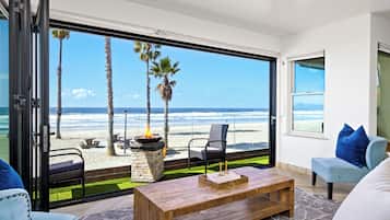 A modern living room with a large glass door leading to a beach view, a wooden coffee table, and blue upholstered chairs.