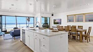A spacious kitchen with a large island, a dining area with wooden chairs, and a view of the ocean through large windows. - Beachfront Bliss - Panoramic Doors & Beach Access - 702-1 (Oceanside)