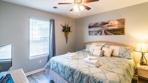 A bedroom with a bed, a ceiling fan, a window with blinds, a wall-mounted picture, and a bedside table with a lamp.