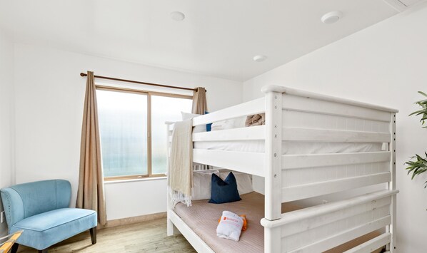 Ein Etagenbett mit weißem Gestell, ein blauer Stuhl und eine Pflanze in einem Zimmer mit großem Fenster.