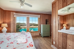 A bedroom with wooden walls, a bed with floral bedding, a green cabinet, and a view of a beach and water through large windows.