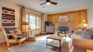 A cozy living room with a fireplace, wooden furniture, and a large window. - Charming Estes Park Cabin w/ 2 Private Patios! (Estes Park)
