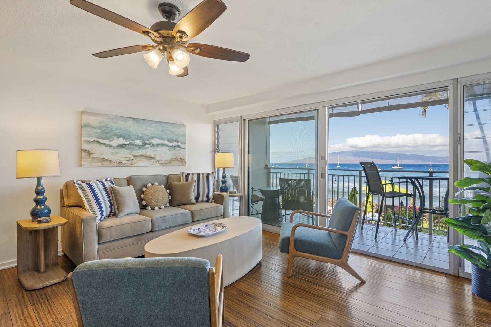 A living room with a sofa, coffee table, and chairs, a ceiling fan, and a balcony with a view of the ocean.