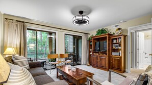 Un salon spacieux avec une table basse en bois, un canapé et une table à manger. Il y a une grande fenêtre avec des rideaux, un ventilateur de plafond et une armoire en bois remplie de livres.