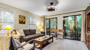 Un salon spacieux avec un canapé, une table basse et des fauteuils, de grandes fenêtres avec des rideaux et une vue sur la verdure environnante.