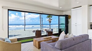 Ein modernes Wohnzimmer mit einem großen Glasfenster, das Blick auf einen Strand bietet, mit einer Couch, Sesseln und einem Couchtisch.