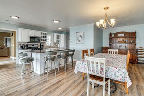 Een moderne keuken met witte kasten, een centraal eiland met barkrukken, een eettafel met stoelen en een houten boekenkast.