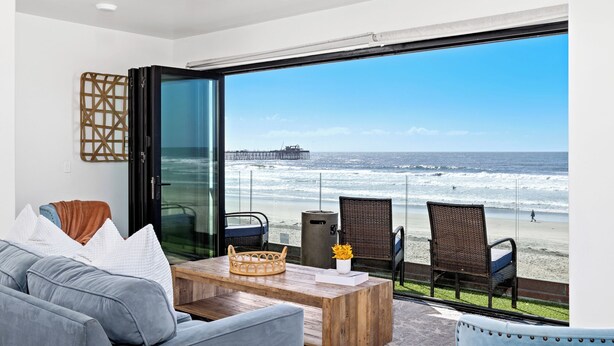 Ein modernes Zimmer mit einem großen Glasfenster mit Blick auf einen Strand, ausgestattet mit einem Holztisch, Korbstühlen und einem grauen Sofa.