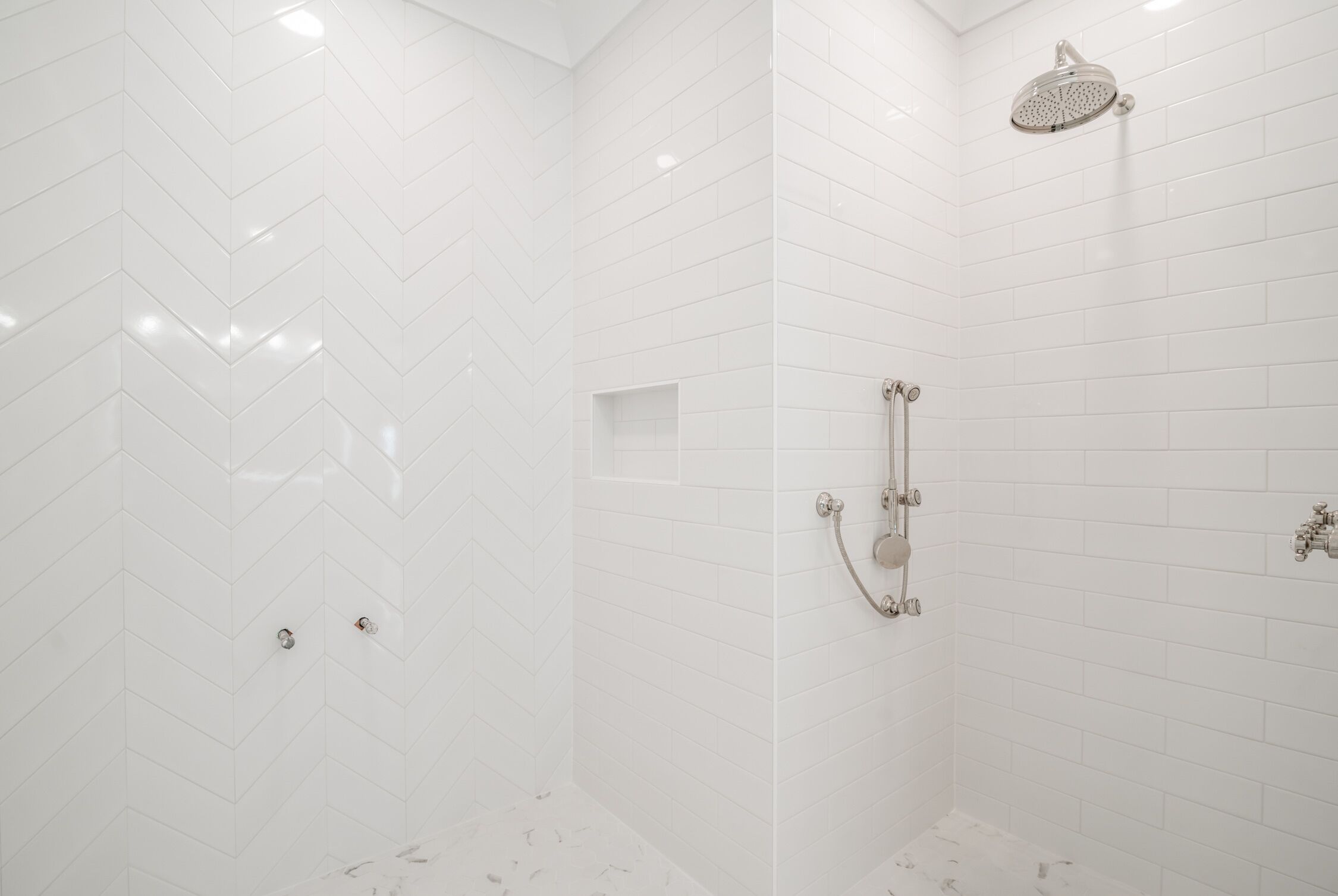 Un baño moderno con azulejos blancos en patrón de espiga, una ducha con cabezal de ducha manual y otro cabezal de ducha manual en la pared.