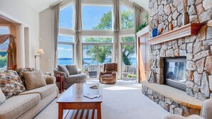 A spacious living room with a stone fireplace, large windows offering a view of the sea, and comfortable seating. - Five Gates: Lakeside Paradise! Seneca Lake Luxury Home! (Geneva)