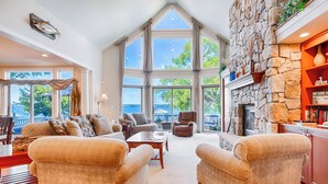 A spacious living room with a stone fireplace, large windows, and a view of the outdoors. - Five Gates: Lakeside Paradise! Seneca Lake Luxury Home! (Geneva)