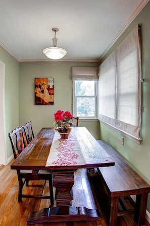 A dining room with a wooden table, chairs, a vase with flowers, a painting on the wall, and a window with blinds. - Vin Rouge 51-Outstanding Westside Paso Robles Home with Tons of Character! (Paso Robles)
